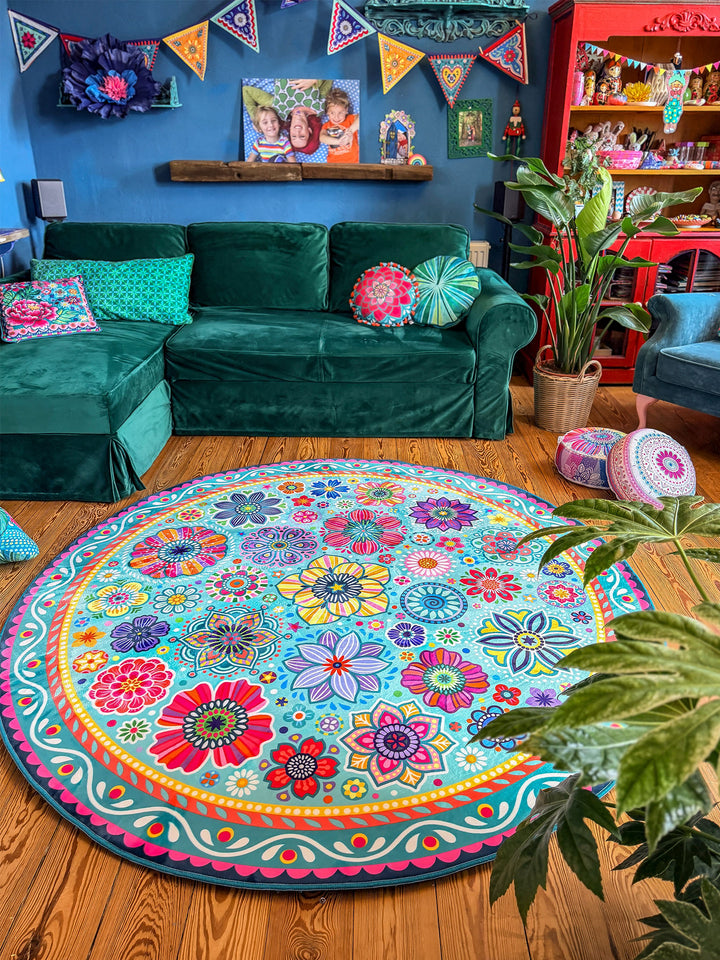 Ein runder Teppich mit buntem Blumen- und Mandala-Muster in Türkis, Pink, Gelb, Rot, Blau und Lila. Sie hebt eine Hand spielerisch nach oben und befindet sich in einem gemütlichen Wohnzimmer mit grünem Samtsofa, Zimmerpflanzen, bunten Kissen und dekorativen Möbeln vor einer dunkelblauen Wand. Der farbenfrohe Teppich bildet den lebendigen Mittelpunkt der kreativen Boho-Wohnatmosphäre.
