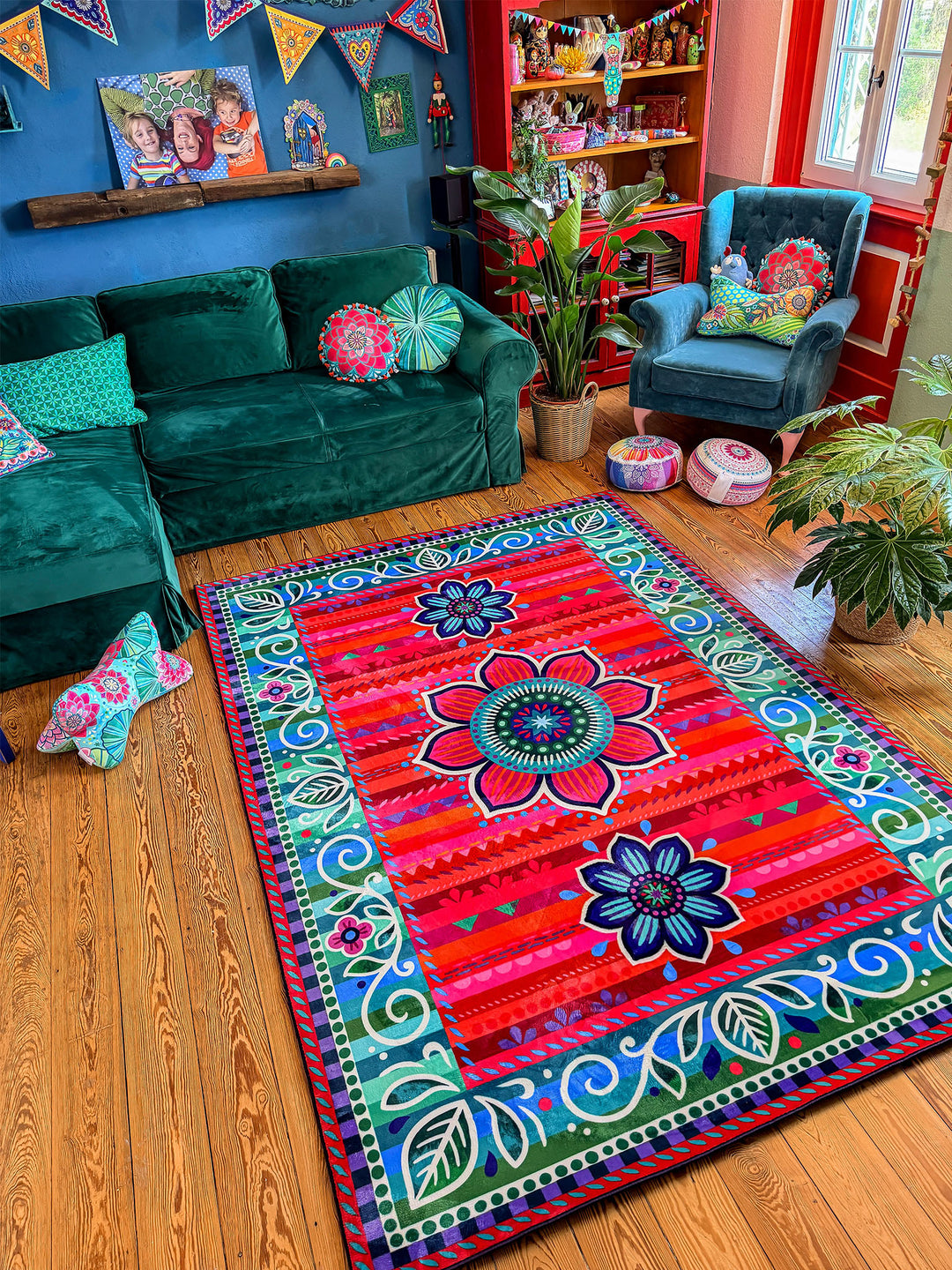 Großer rechteckiger Teppich mit farbenfrohem Blumen- und Ornamentmuster in Rot, Pink, Türkis, Blau, Grün und Lila liegt auf warmem Holzboden in einem gemütlichen Wohnzimmer. Im Hintergrund stehen ein grünes Samtsofa, ein blauer Sessel, Zimmerpflanzen und dekorative Möbel vor einer dunkelblauen Wand mit bunten Girlanden. Der bunte Teppich setzt einen lebendigen Mittelpunkt und schafft eine kreative, wohnliche Boho-Atmosphäre.