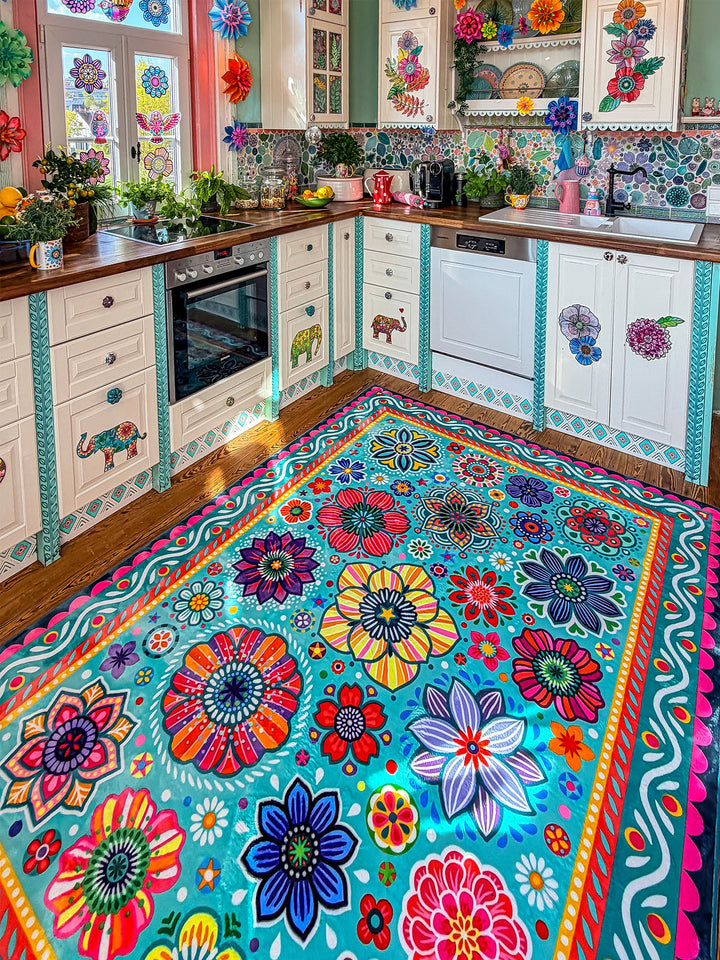 Großer Teppich in Türkis mit farbenfrohen Blumen- und Mandala-Mustern in Pink, Gelb, Rot, Blau, Grün und Lila liegt auf Holzboden in einer kreativ gestalteten Küche. Im Hintergrund stehen helle Küchenschränke mit dekorativen Elefantenmotiven, Holzarbeitsplatten, Pflanzen, Obstschalen und bunte Fensterdekorationen. Der florale Teppich bringt lebendige Farben, gemütliche Wärme und verspielten Boho-Stil in den Küchenbereich.