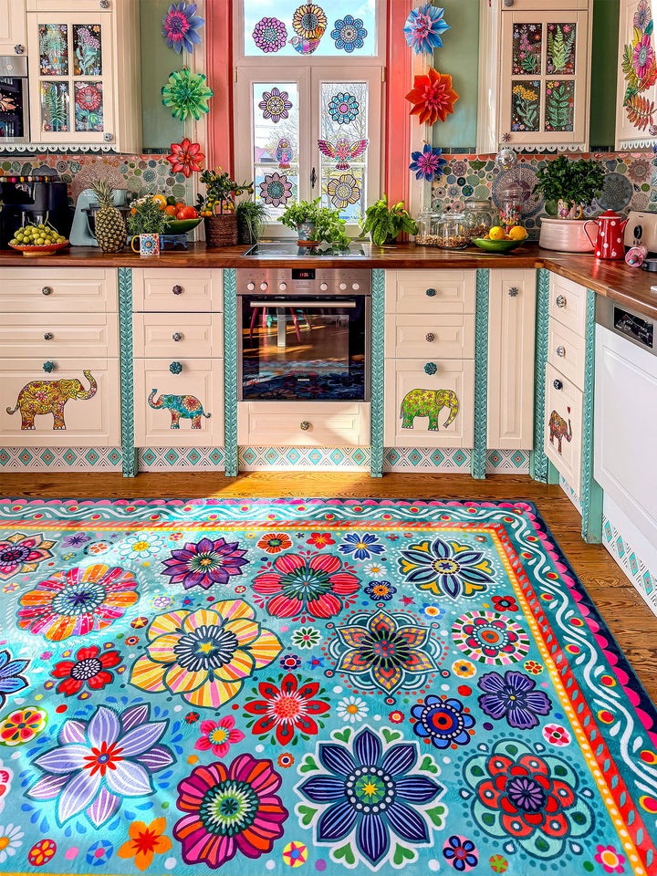 Großer Teppich in Türkis mit farbenfrohen Blumen- und Mandala-Mustern in Pink, Gelb, Rot, Blau, Grün und Lila liegt auf Holzboden in einer kreativ gestalteten Küche. Im Hintergrund stehen helle Küchenschränke mit dekorativen Elefantenmotiven, Holzarbeitsplatten, Pflanzen, Obstschalen und bunte Fensterdekorationen. Der florale Teppich bringt lebendige Farben, gemütliche Wärme und verspielten Boho-Stil in den Küchenbereich.