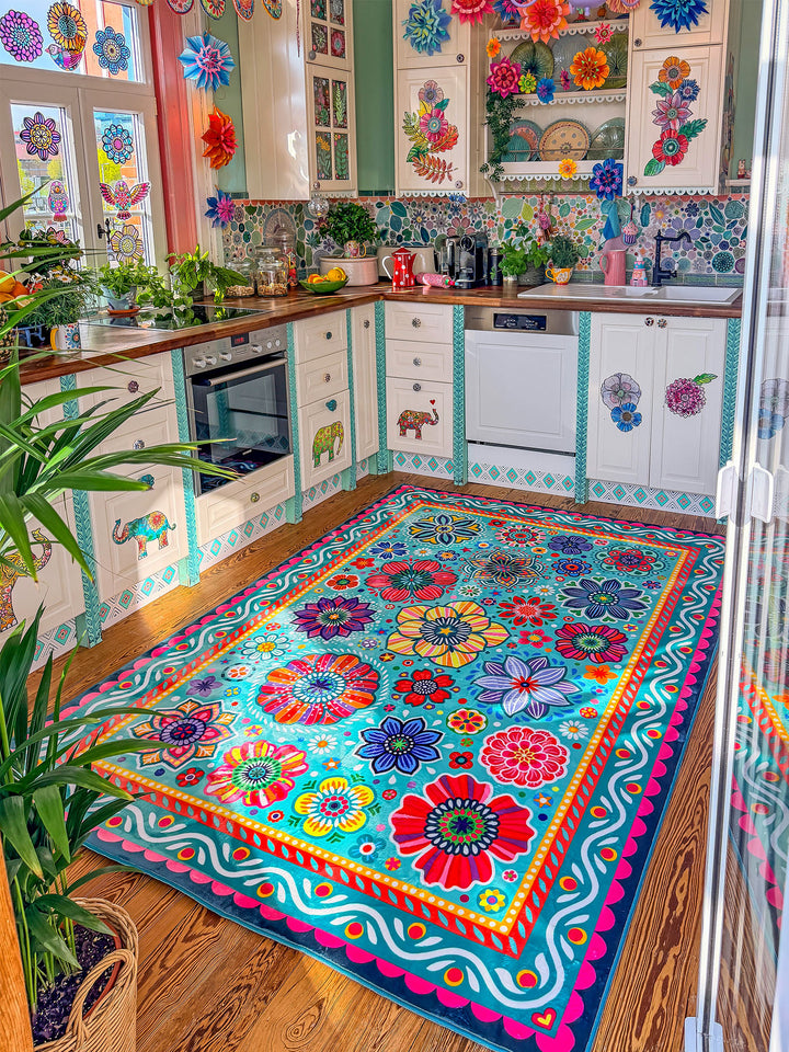 Großer Teppich in Türkis mit farbenfrohen Blumen- und Mandala-Mustern in Pink, Gelb, Rot, Blau, Grün und Lila liegt auf Holzboden in einer kreativ gestalteten Küche. Im Hintergrund stehen helle Küchenschränke mit dekorativen Elefantenmotiven, Holzarbeitsplatten, Pflanzen, Obstschalen und bunte Fensterdekorationen. Der florale Teppich bringt lebendige Farben, gemütliche Wärme und verspielten Boho-Stil in den Küchenbereich.