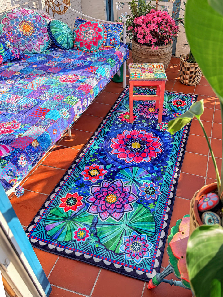 Langer Läuferteppich mit floralem Mandala-Muster in Dunkelblau, Pink, Lila, Türkis und Grün liegt auf terrakottafarbenen Fliesen in einem hellen Wintergarten oder Balkon. Daneben stehen eine bunt bezogene Sitzbank, dekorative Kissen, ein kleiner rosa Hocker und blühende Pflanzen. Große grüne Blätter rahmen die Szene ein. Der farbenfrohe Teppich schafft eine gemütliche, lebendige Boho-Wohnatmosphäre mit mediterranem Flair.