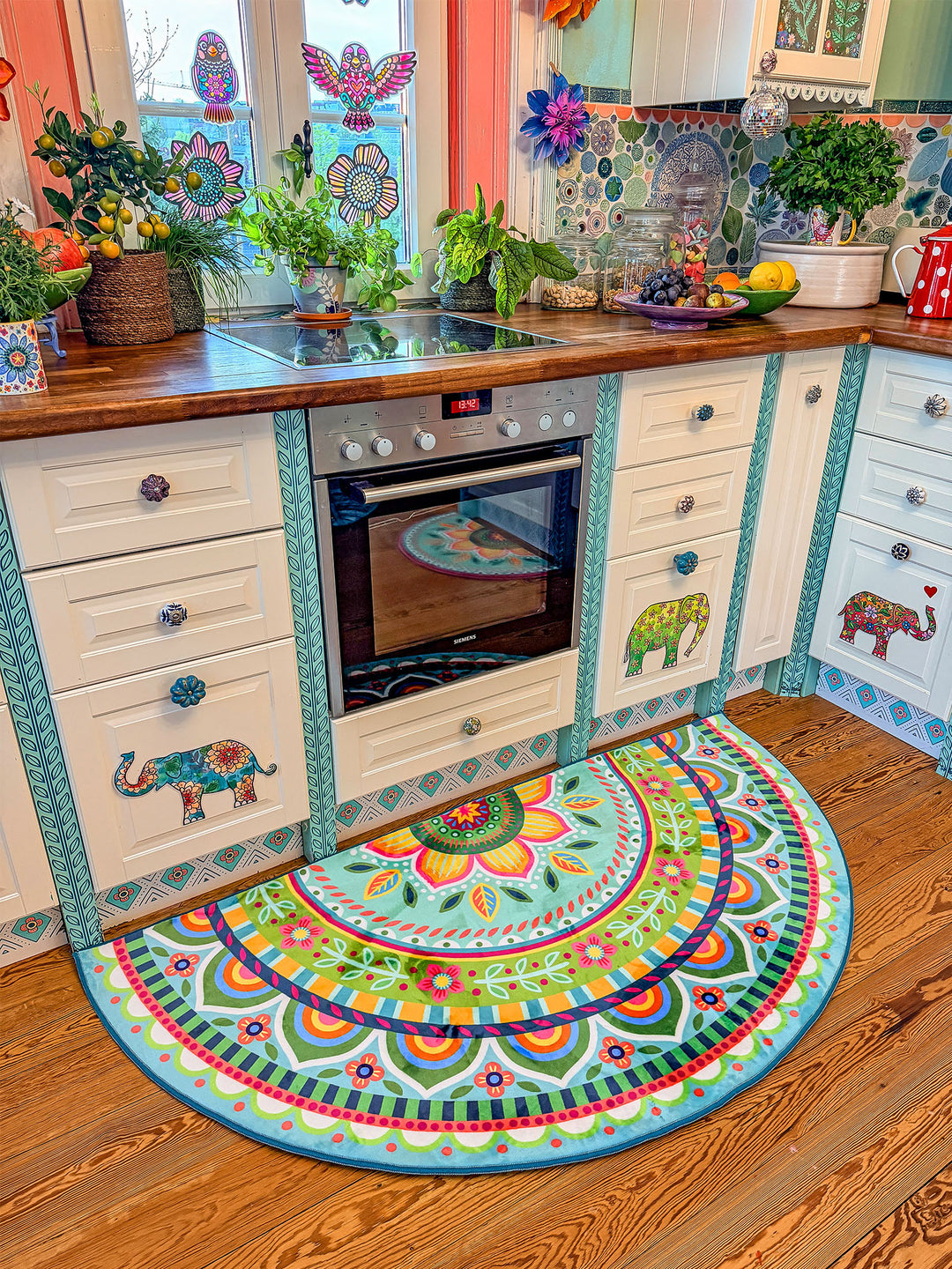 Halbrunder Küchenteppich in Pastell-Türkis mit detailreichen Mandala- und Blumenmustern in Pink, Gelb, Grün, Orange und Blau liegt vor einer hellen Landhausküche mit Holzarbeitsplatte. Die weißen Schränke sind mit farbenfrohen Elefantenmotiven verziert. Über dem Spülbecken befindet sich ein dekoriertes Fenster mit bunten Blumenstickern und Papierblüten. Pflanzen, Obstschalen und liebevolle Details schaffen eine warme, kreative Boho-Atmosphäre.