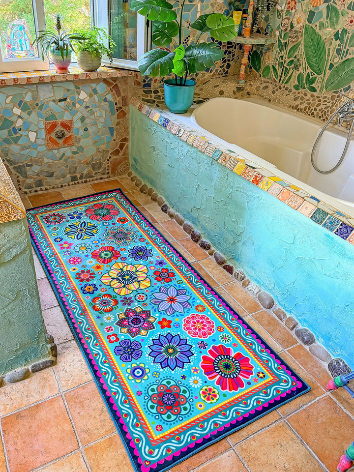 Schmaler rechteckiger Badteppich in Türkis mit farbenfrohen Blumen- und Mandalamotiven in Pink, Blau, Gelb, Rot und Grün liegt auf terrakottafarbenen Fliesen vor einer Badewanne. Das kreative Badezimmer ist mit kunstvollen Mosaikwänden, Naturstein-Details und Pflanzen dekoriert. Große grüne Blätter und ein Fenster mit Tageslicht schaffen eine frische, natürliche Atmosphäre. Der lebendige Teppich setzt einen fröhlichen Farbakzent im individuell gestalteten Boho-Bad.