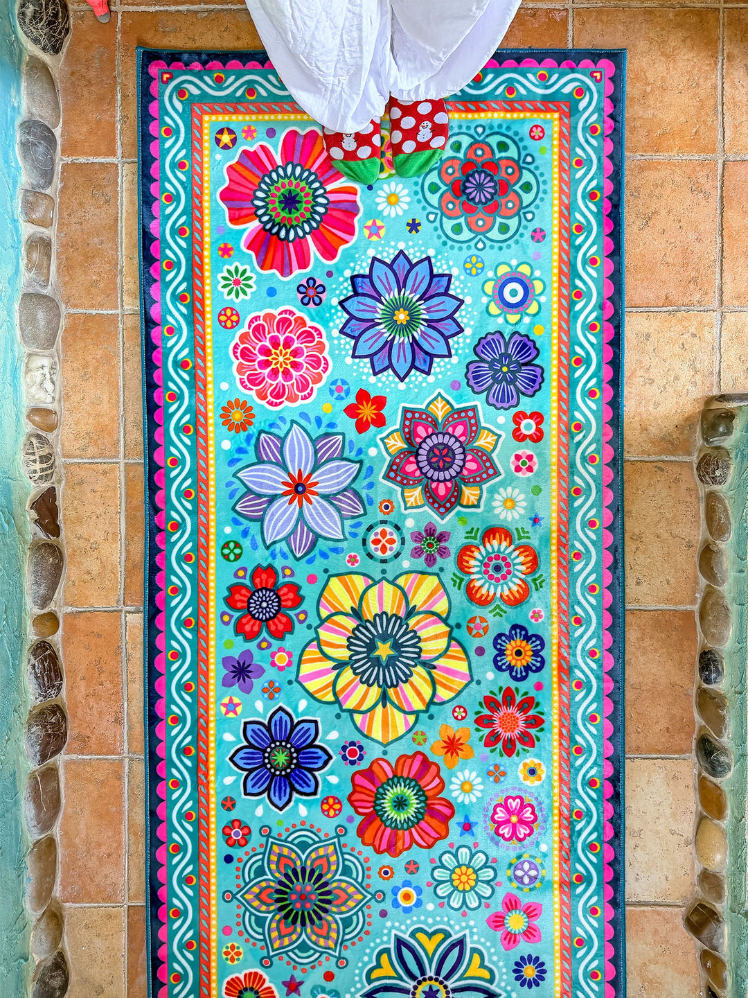 Schmaler rechteckiger Badteppich in Türkis mit farbenfrohen Blumen- und Mandalamotiven in Pink, Blau, Gelb, Rot und Grün liegt auf terrakottafarbenen Fliesen vor einer Badewanne. Das kreative Badezimmer ist mit kunstvollen Mosaikwänden, Naturstein-Details und Pflanzen dekoriert. Große grüne Blätter und ein Fenster mit Tageslicht schaffen eine frische, natürliche Atmosphäre. Der lebendige Teppich setzt einen fröhlichen Farbakzent im individuell gestalteten Boho-Bad.