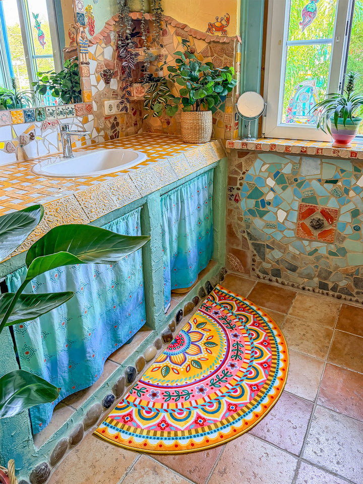 Halbrunder Badteppich in leuchtendem Gelb mit farbenfrohem Mandala- und Blumenmuster in Pink, Türkis, Orange, Blau und Grün liegt auf warmen Natursteinfliesen vor einem Waschtisch. Der rustikal-kreative Waschtisch besitzt eine gelb-weiß geflieste Ablage, ein weißes Waschbecken und türkisfarbene Stoffvorhänge darunter. Naturstein- und Mosaikdetails an Wand und Möbeln verleihen dem Badezimmer einen kunstvollen Boho-Stil. Die fröhliche Matte setzt einen lebendigen Farbakzent im Raum.