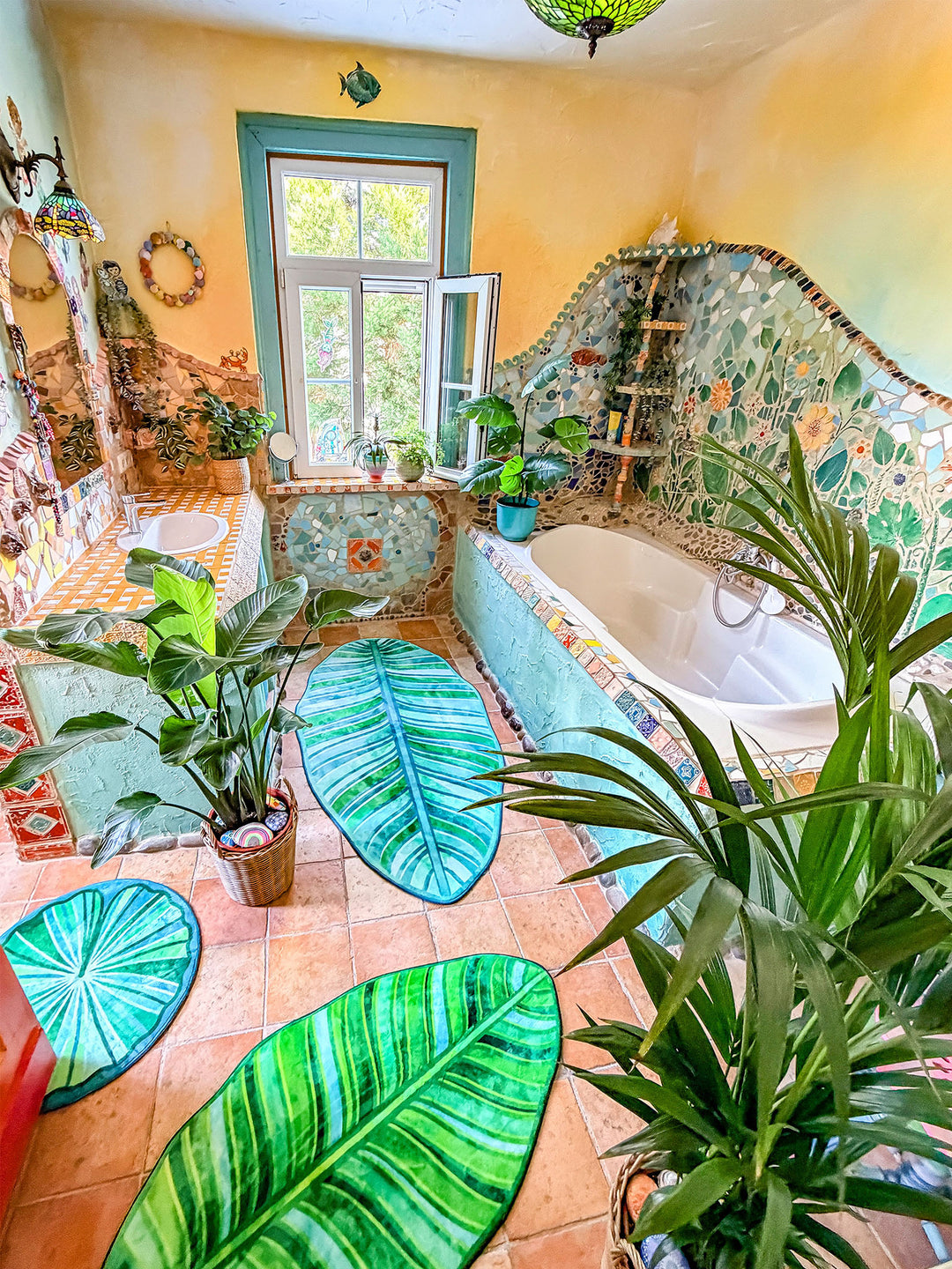 Künstlerisch gestaltetes Badezimmer im Boho- und Tropenstil mit mehreren grünen Badteppichen in Blattform auf terrakottafarbenen Fliesen. Die Teppiche erinnern an große tropische Blätter mit detailreichen Blattadern in verschiedenen Grün- und Türkistönen. Im Mittelpunkt steht eine große Zimmerpflanze im Korb, umgeben von den Teppichen. Der Waschtisch ist mit gelb-weißen Mosaikfliesen verkleidet und besitzt türkisfarbene Stoffvorhänge als Stauraum. Wände, Fensterrahmen und Badewanne sind mit Naturstein, bunt
