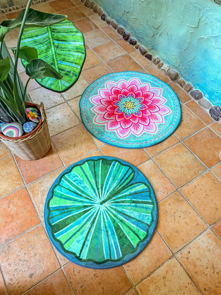 Drei dekorative Badteppiche liegen auf warmen Terrakottafliesen in einem kreativen Badezimmer im Boho-Stil. Oben links befindet sich ein großer grüner Teppich in Form eines tropischen Blattes mit detailreichen Blattadern. In der Mitte liegt ein runder Teppich in Türkis mit großer pinker Blüte im Mandala-Stil. Im Vordergrund ergänzt ein weiterer runder Teppich in Grün- und Türkistönen, inspiriert von einem Seerosenblatt, das Arrangement. Links steht eine große Zimmerpflanze im geflochtenen Korb mit bunt bema
