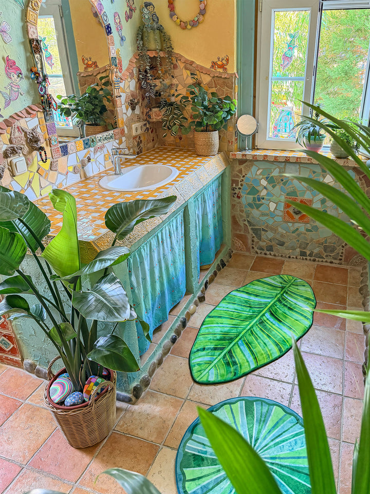 Farbenfrohes Badezimmer im Boho-Stil mit mehreren dekorativen Badteppichen auf warmen Terrakottafliesen. Im Vordergrund liegt ein großer Teppich in Form eines tropischen Blattes mit detailreichen Blattadern in satten Grün- und Limetönen. Dahinter ergänzen ein runder grüner Blätterteppich und ein halbrunder Mandala-Teppich in Türkis, Pink, Gelb und Blau das harmonische Gesamtbild. Links steht eine große Zimmerpflanze im geflochtenen Korb, rechts befindet sich eine Badewanne mit türkis verputzter Front und bu