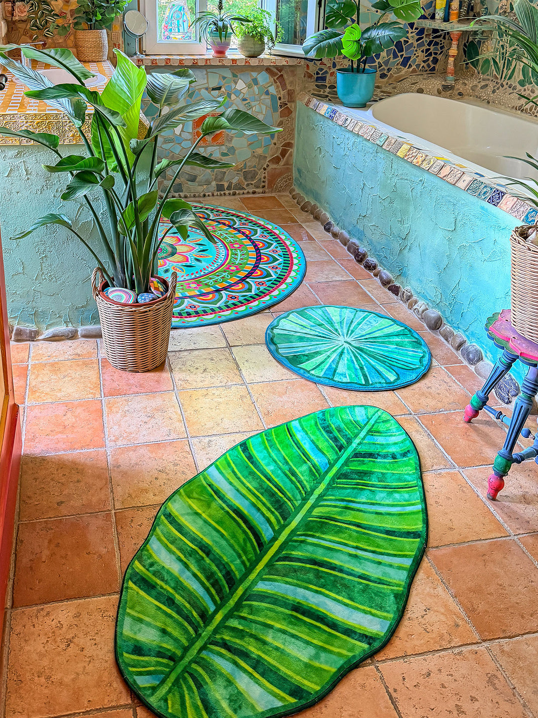 Farbenfrohes Badezimmer im Boho-Stil mit mehreren dekorativen Badteppichen auf warmen Terrakottafliesen. Im Vordergrund liegt ein großer Teppich in Form eines tropischen Blattes mit detailreichen Blattadern in satten Grün- und Limetönen. Dahinter ergänzen ein runder grüner Blätterteppich und ein halbrunder Mandala-Teppich in Türkis, Pink, Gelb und Blau das harmonische Gesamtbild. Links steht eine große Zimmerpflanze im geflochtenen Korb, rechts befindet sich eine Badewanne mit türkis verputzter Front und bu