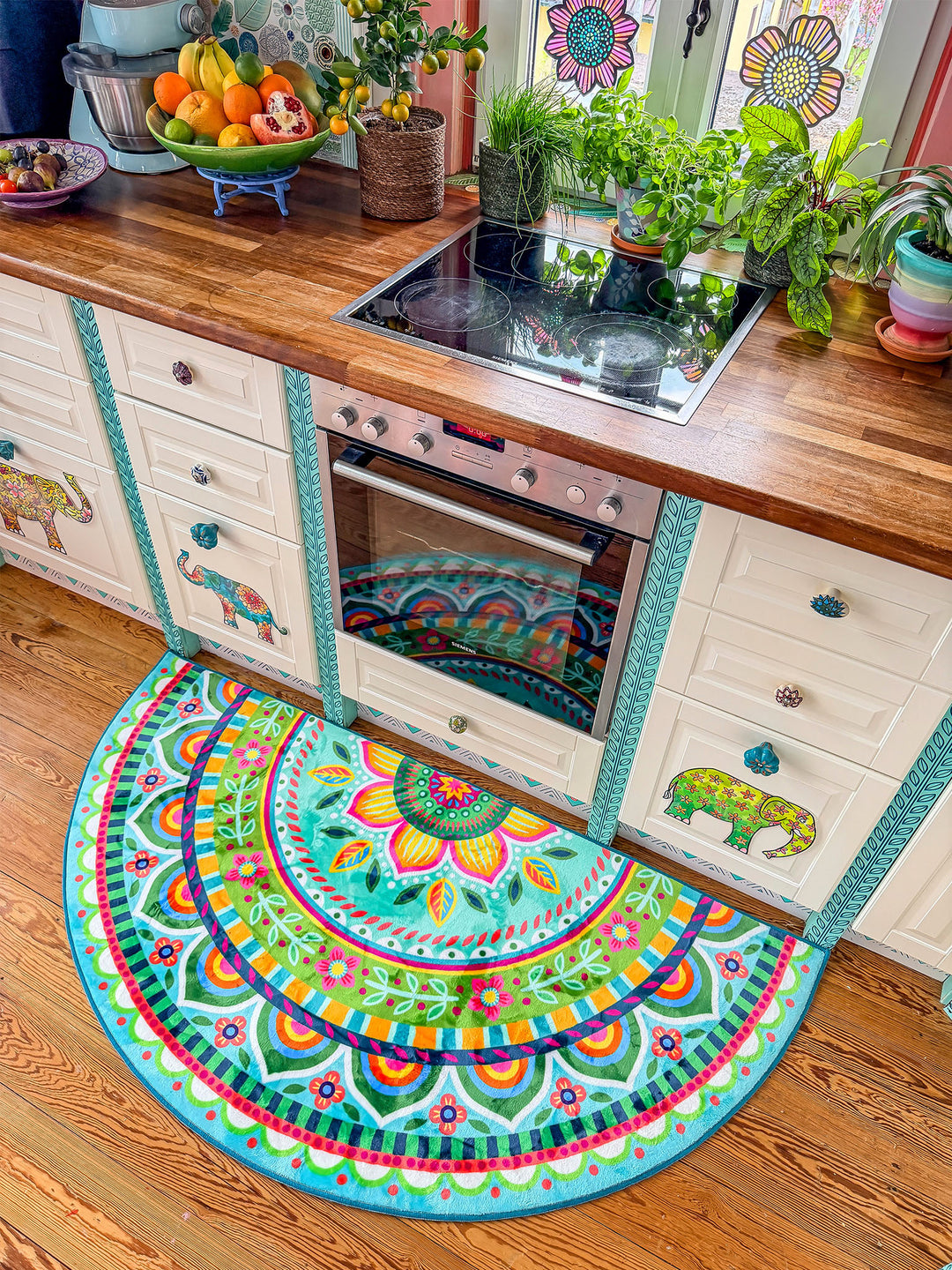 Halbrunder Küchenteppich in Türkis mit farbenfrohem Mandala- und Blumenmuster liegt vor einem Backofen auf hellem Holzboden. Die kreative Landhausküche besitzt weiße Schränke mit dekorativen Tiermotiven, Holzarbeitsplatte, Cerankochfeld sowie viele grüne Kräuter und Zimmerpflanzen am Fenster. Obstschale, bunte Details und verspielte Muster schaffen eine gemütliche, lebendige Küchenatmosphäre.