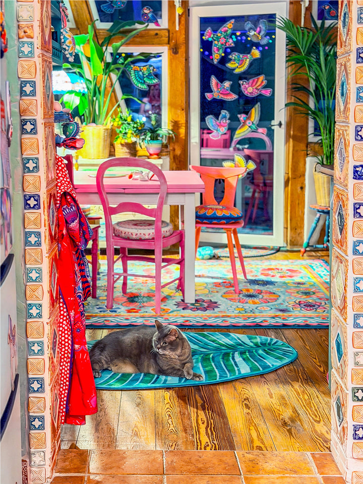 Graue Katze liegt entspannt auf einem grünen Blatt-Teppich im Flur eines farbenfrohen Wohnraums. Im Hintergrund stehen ein rosa Tisch mit bunten Stühlen, Zimmerpflanzen und ein großer Teppich mit Blumenmustern. Die Glastür ist mit dekorativen Vogelmotiven gestaltet. Holzfußboden, warme Farben und kreative Einrichtung schaffen eine gemütliche, verspielte Wohnatmosphäre.